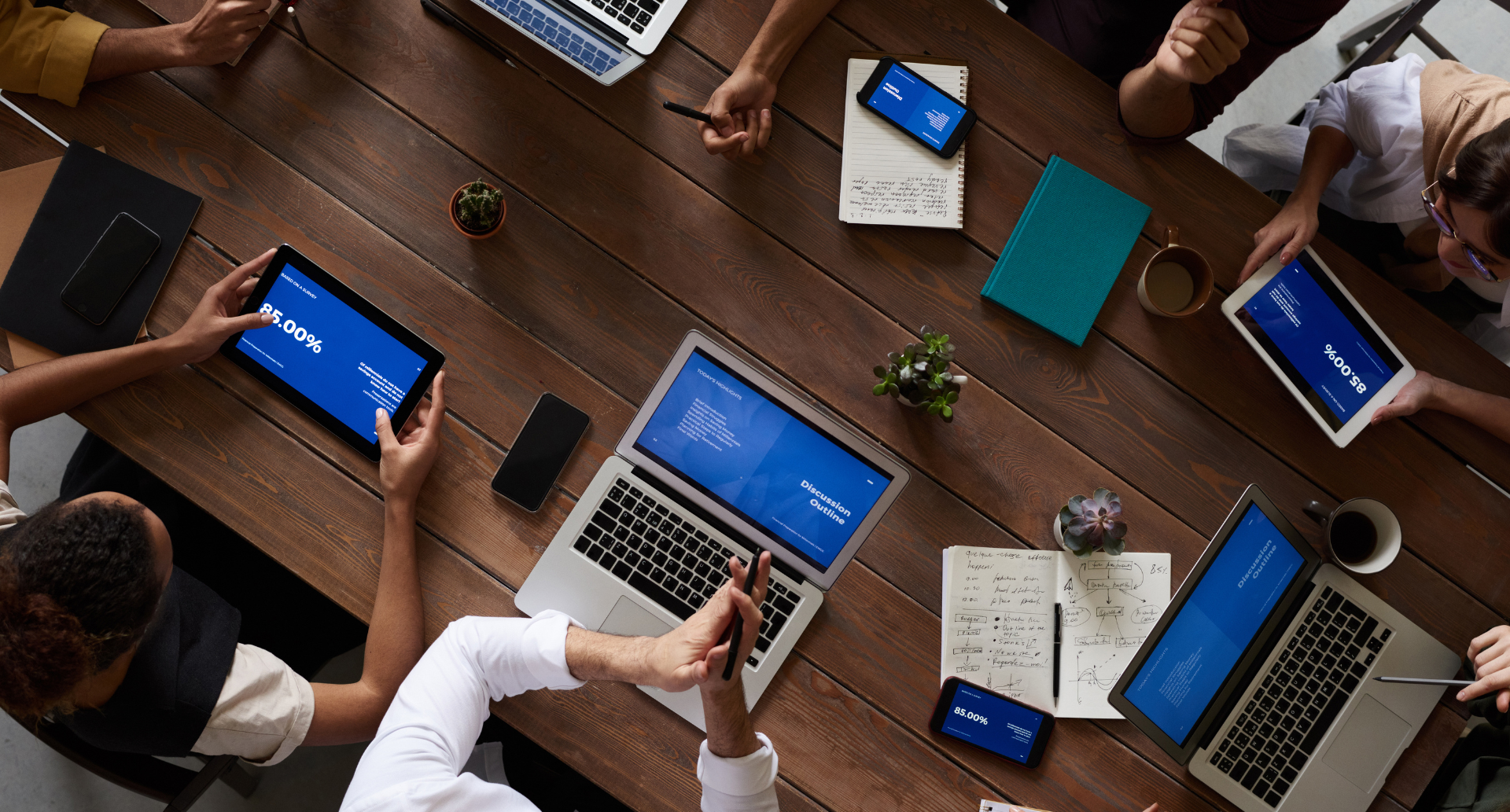Business meeting overhead showing team using laptops and tablets at a wooden table.